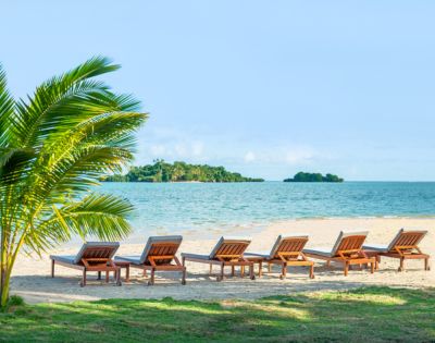Sun-drenched beach with six empty wooden lounge chairs facing the turquoise ocean. A lush palm tree on the left adds a tropical feel. Relaxing and serene.