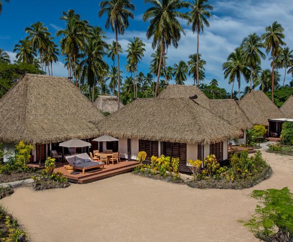 Thatched-roof bungalows surrounded by palm trees on a sandy beach, with lounge chairs under umbrellas on wooden decks, evoke a tropical paradise feel.