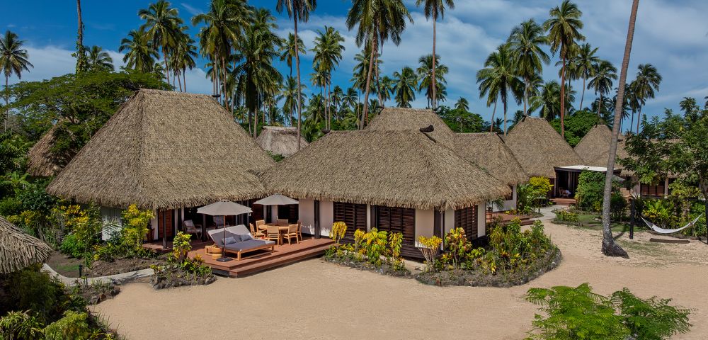 Thatched-roof bungalows surrounded by palm trees on a sandy beach, with lounge chairs under umbrellas on wooden decks, evoke a tropical paradise feel.