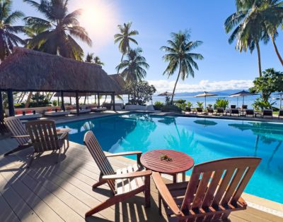 Luxurious tropical poolside with palm trees, thatched-roof pavilion, and wooden lounge chairs. The scene is serene, sunny, and inviting.
