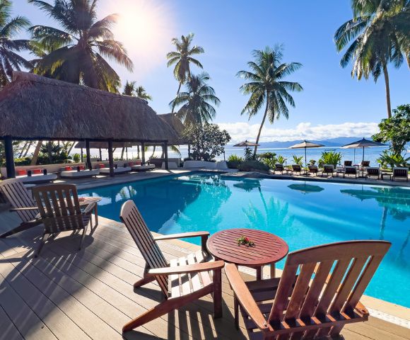 Luxurious tropical poolside with palm trees, thatched-roof pavilion, and wooden lounge chairs. The scene is serene, sunny, and inviting.