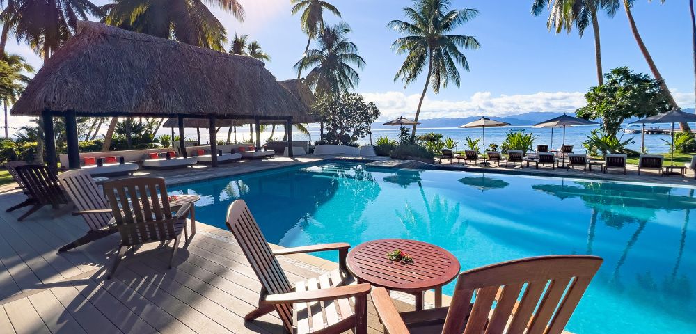 Luxurious tropical poolside with palm trees, thatched-roof pavilion, and wooden lounge chairs. The scene is serene, sunny, and inviting.