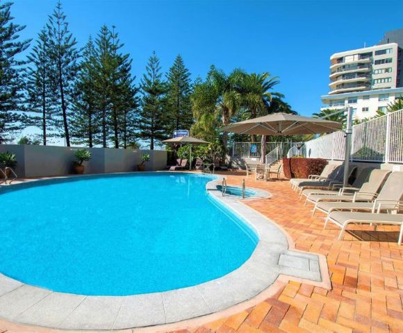 Sunny outdoor swimming pool area with clear blue water, surrounded by red brick patio, lounge chairs, and umbrellas. Tall trees and modern buildings in the background. Relaxing and inviting atmosphere.