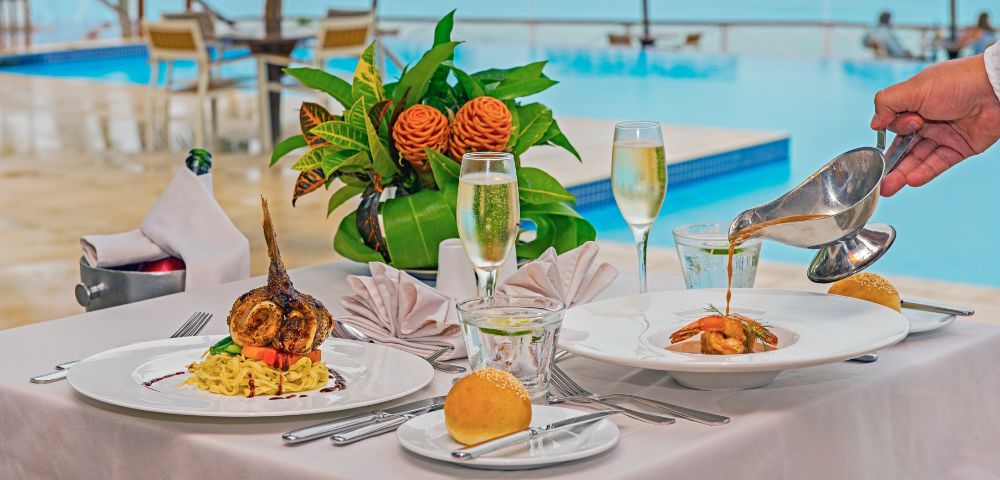 Elegant poolside dining setup with gourmet dishes, fresh floral centerpiece, champagne glasses, and a bread roll, exuding luxury and relaxation.
