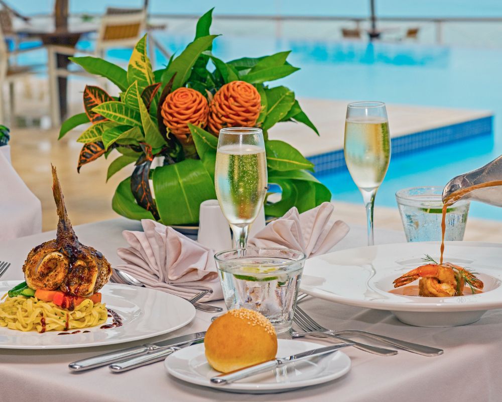 Elegant poolside dining setup with gourmet dishes, fresh floral centerpiece, champagne glasses, and a bread roll, exuding luxury and relaxation.