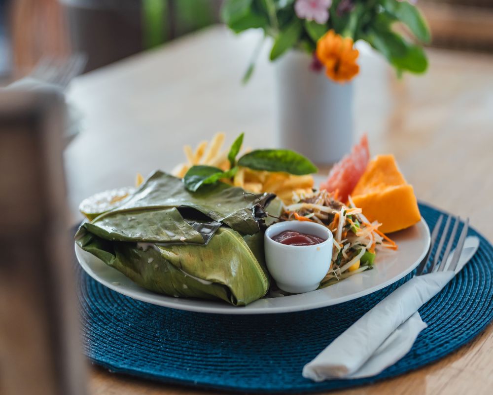 A plate with food wrapped in banana leaves, accompanied by fries, a small salad, melon slices, and a sauce cup. The setting is bright and inviting.