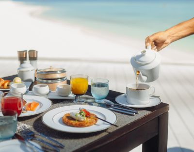 A luxurious breakfast setup on a table by the beach, featuring pastries, juice, and a hand pouring tea into a cup, evoking a serene, indulgent vibe.
