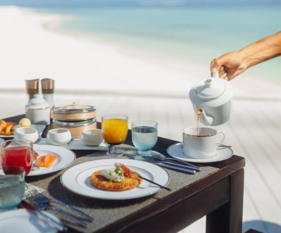 A luxurious breakfast setup on a table by the beach, featuring pastries, juice, and a hand pouring tea into a cup, evoking a serene, indulgent vibe.