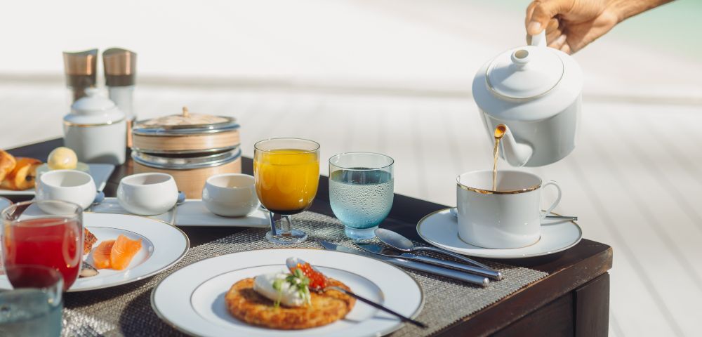 A luxurious breakfast setup on a table by the beach, featuring pastries, juice, and a hand pouring tea into a cup, evoking a serene, indulgent vibe.