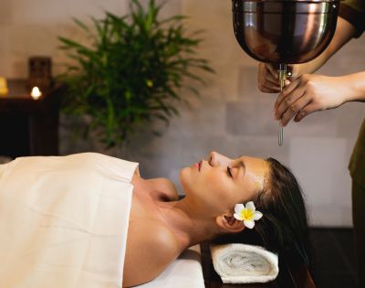 A woman receiving a relaxing oil massage at a spa, lying with eyes closed and a flower in her hair. The atmosphere is tranquil, with a plant and soft lighting.