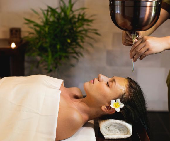 A woman receiving a relaxing oil massage at a spa, lying with eyes closed and a flower in her hair. The atmosphere is tranquil, with a plant and soft lighting.