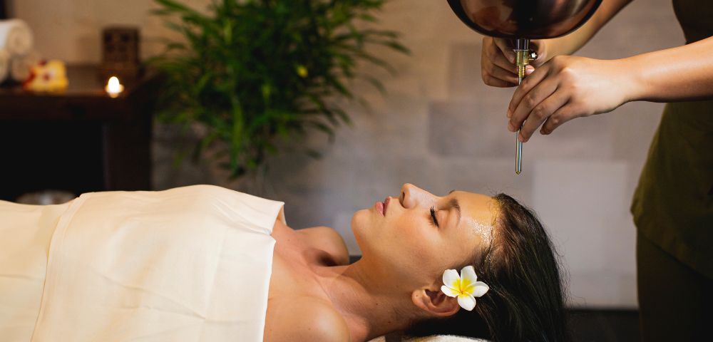 A woman receiving a relaxing oil massage at a spa, lying with eyes closed and a flower in her hair. The atmosphere is tranquil, with a plant and soft lighting.
