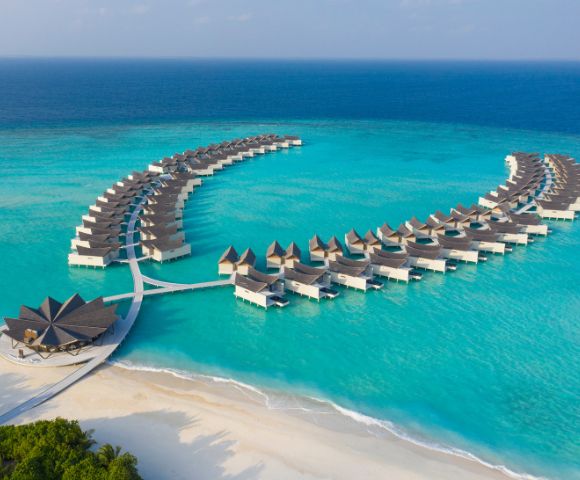 Luxurious overwater bungalows shaped in two arcs rest on turquoise ocean waters. A sandy beach with lush greenery completes the tranquil tropical scene.