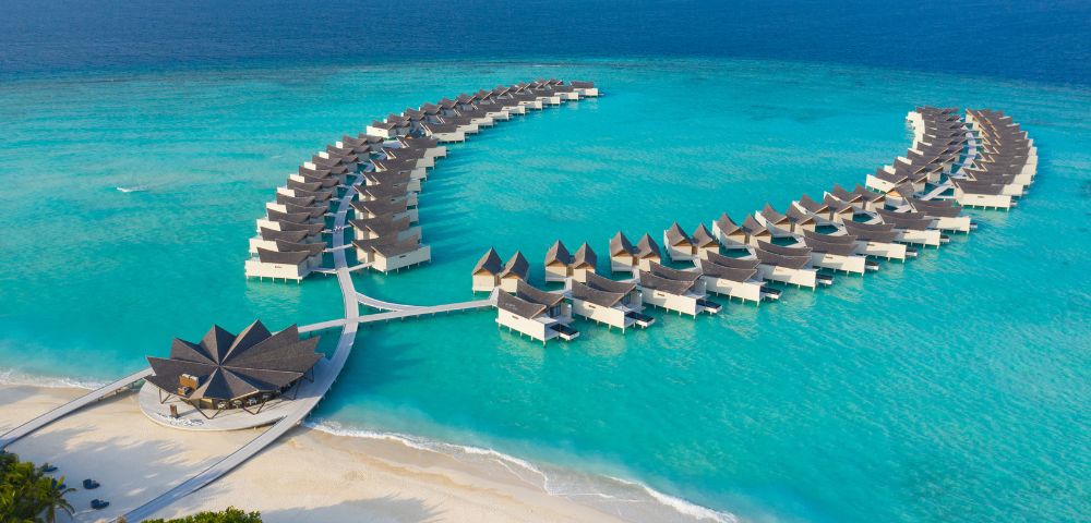 Luxurious overwater bungalows shaped in two arcs rest on turquoise ocean waters. A sandy beach with lush greenery completes the tranquil tropical scene.