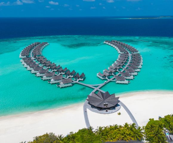 Aerial view of a tropical resort with overwater bungalows arranged symmetrically in turquoise waters, white sandy beach, under a clear blue sky.