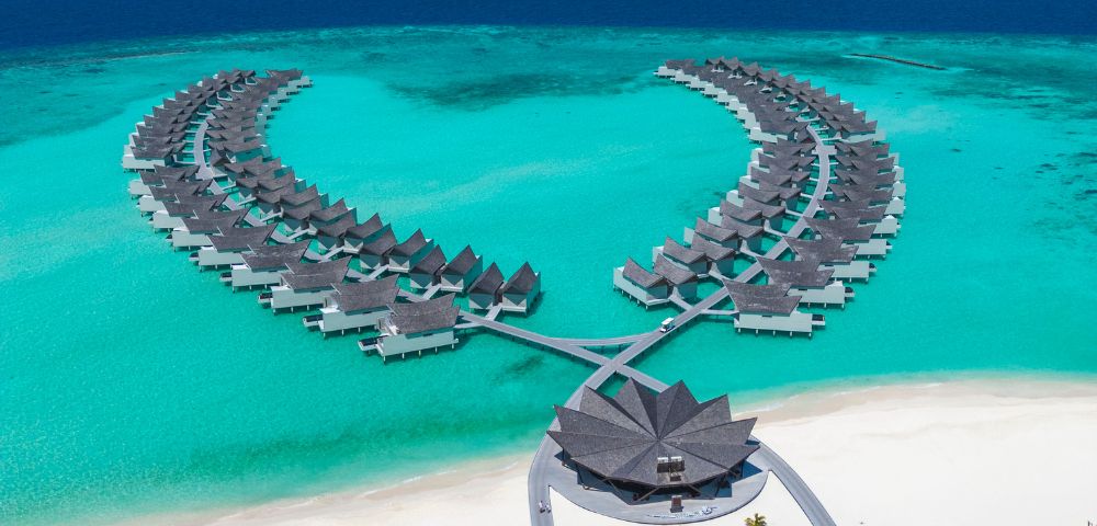 Aerial view of a tropical resort with overwater bungalows arranged symmetrically in turquoise waters, white sandy beach, under a clear blue sky.