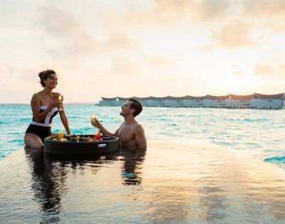A couple enjoys drinks by an infinity pool at sunset, with the ocean and overwater bungalows in the background, conveying a serene, tropical getaway vibe.