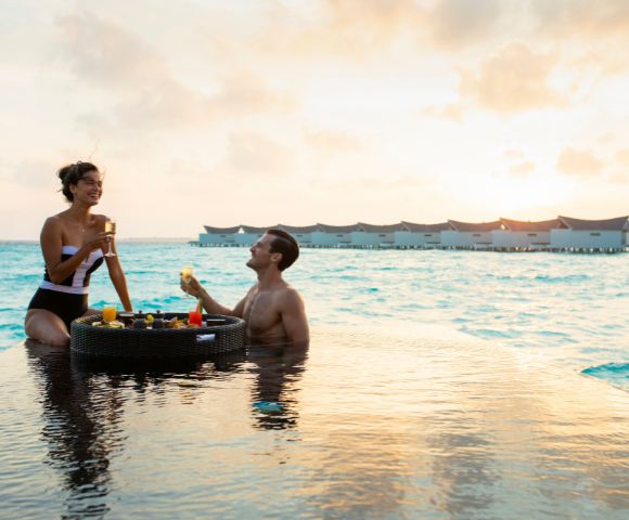 A couple enjoys drinks by an infinity pool at sunset, with the ocean and overwater bungalows in the background, conveying a serene, tropical getaway vibe.