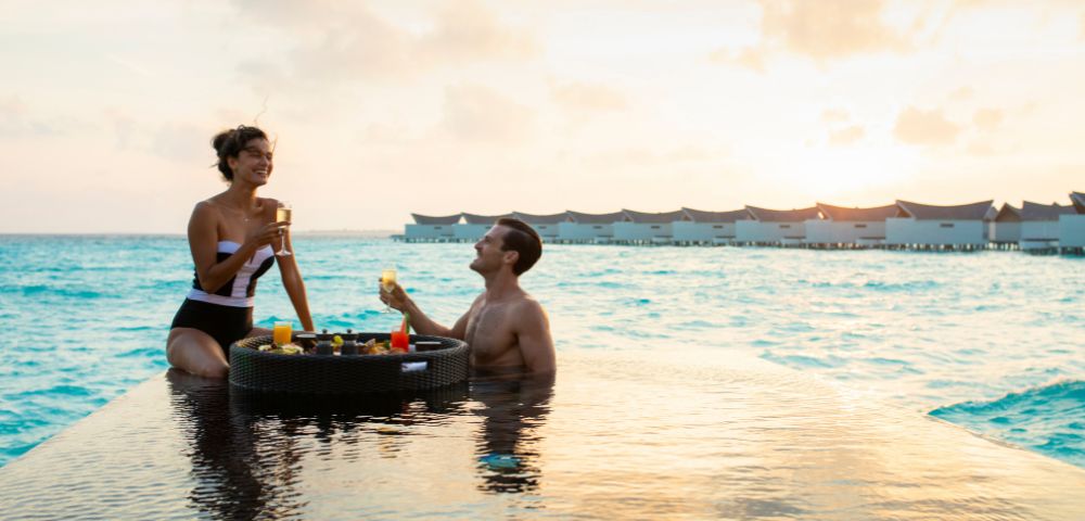 A couple enjoys drinks by an infinity pool at sunset, with the ocean and overwater bungalows in the background, conveying a serene, tropical getaway vibe.