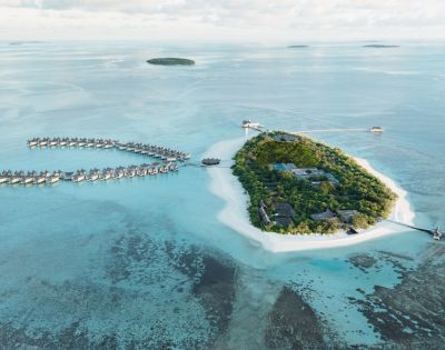 Aerial view of tropical island resort with lush greenery, white sand beaches, and overwater bungalows in turquoise water, under a vast blue sky.
