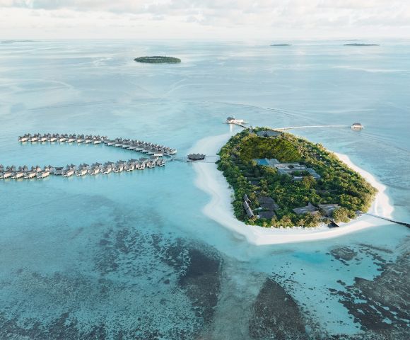 Aerial view of tropical island resort with lush greenery, white sand beaches, and overwater bungalows in turquoise water, under a vast blue sky.