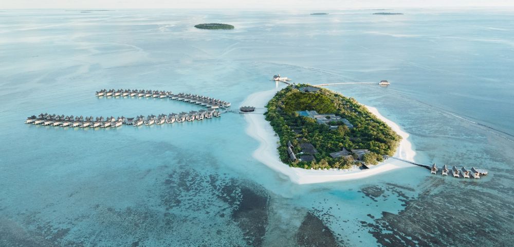 Aerial view of tropical island resort with lush greenery, white sand beaches, and overwater bungalows in turquoise water, under a vast blue sky.