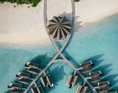 Aerial view of a tropical resort with thatched-roof villas on turquoise water. Villas are connected by walkways, creating a symmetrical pattern. Serene and luxurious.