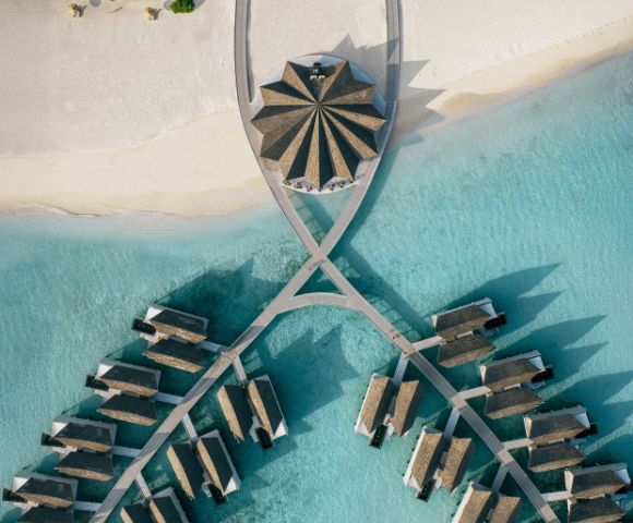 Aerial view of a tropical resort with thatched-roof villas on turquoise water. Villas are connected by walkways, creating a symmetrical pattern. Serene and luxurious.