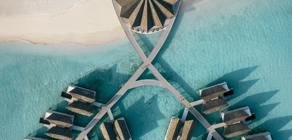 Aerial view of a tropical resort with thatched-roof villas on turquoise water. Villas are connected by walkways, creating a symmetrical pattern. Serene and luxurious.