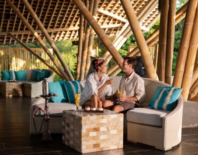 A couple sits smiling on a wicker sofa in an open-air bamboo lounge, holding drinks. The setting is cozy, relaxed, with teal cushions and warm lighting.