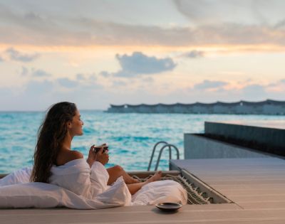 A woman in a white robe reclines on a deck overlooking a serene ocean at sunset, holding a cup. The scene conveys relaxation and tranquility.