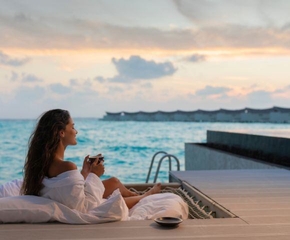 A woman in a white robe reclines on a deck overlooking a serene ocean at sunset, holding a cup. The scene conveys relaxation and tranquility.