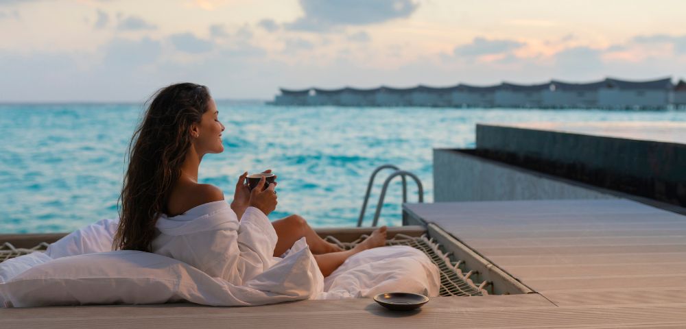 A woman in a white robe reclines on a deck overlooking a serene ocean at sunset, holding a cup. The scene conveys relaxation and tranquility.