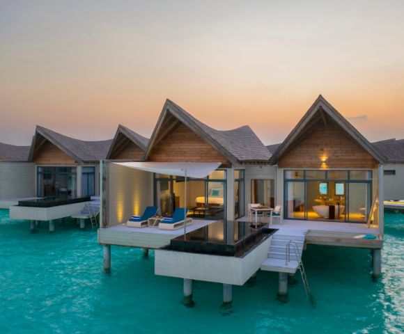 Overwater bungalows on a serene turquoise sea at sunset. Each has a private deck, plunge pool, and lounge chairs, creating a tranquil, luxurious vibe.