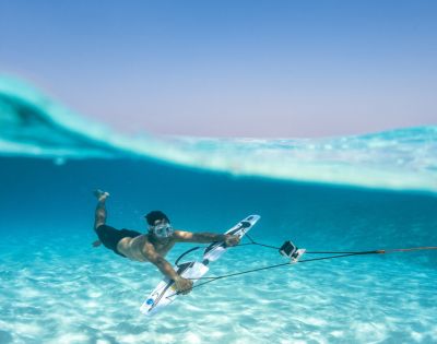 A man underwater in clear blue sea, wearing goggles and holding a board with a mounted camera, conveys adventure and exploration in a serene setting.
