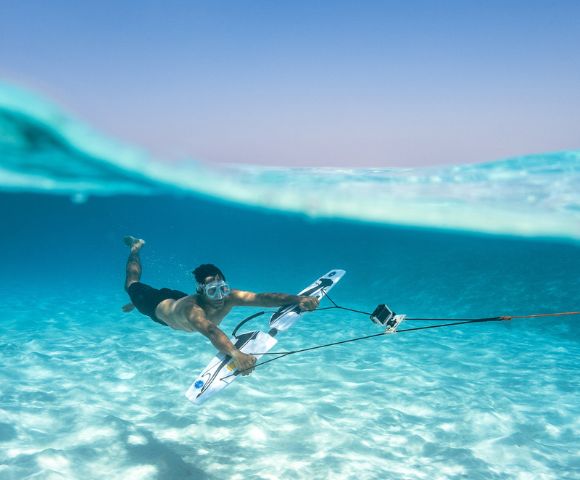 A man underwater in clear blue sea, wearing goggles and holding a board with a mounted camera, conveys adventure and exploration in a serene setting.