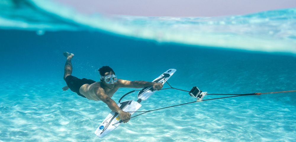 A man underwater in clear blue sea, wearing goggles and holding a board with a mounted camera, conveys adventure and exploration in a serene setting.