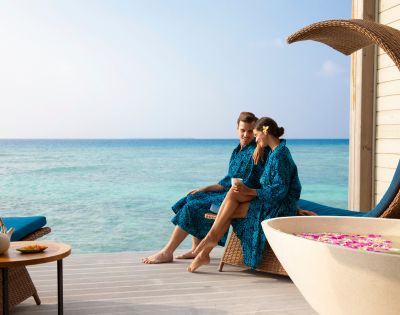 A couple in blue robes relaxes on a deck by the ocean, sitting on a wicker lounge chair. A teapot is nearby, and a tub with flower petals adds to the serene ambiance.
