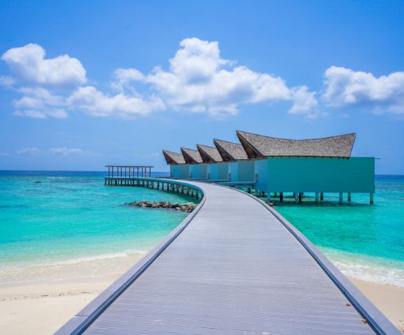Curved boardwalk leads to overwater villas with slanted roofs, surrounded by turquoise sea under a bright blue sky with white clouds; serene and tropical.