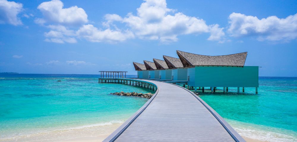 Curved boardwalk leads to overwater villas with slanted roofs, surrounded by turquoise sea under a bright blue sky with white clouds; serene and tropical.