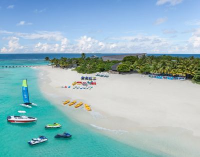 A tropical beach with white sand, turquoise waters, and anchored boats. Vibrant kayaks and parasailing gear line the shore, evoking a lively, adventurous vibe.