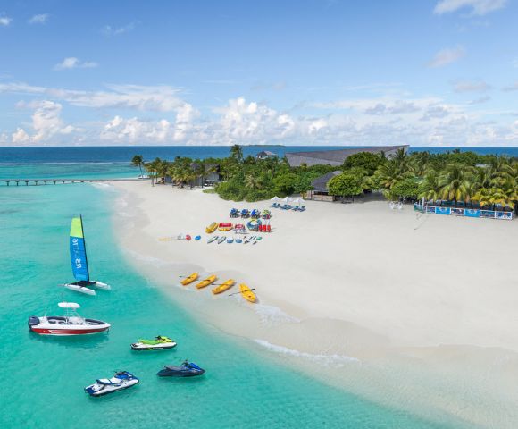 A tropical beach with white sand, turquoise waters, and anchored boats. Vibrant kayaks and parasailing gear line the shore, evoking a lively, adventurous vibe.