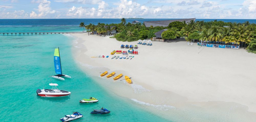 A tropical beach with white sand, turquoise waters, and anchored boats. Vibrant kayaks and parasailing gear line the shore, evoking a lively, adventurous vibe.