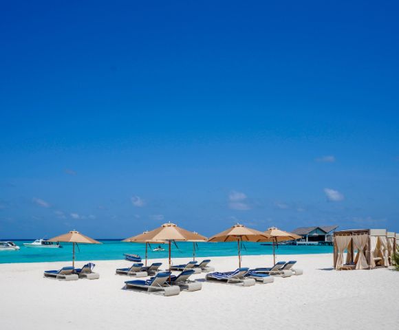 Sunlit beach with blue waters, beige umbrellas, and lounge chairs. Boats float on the horizon, and cabanas line the right side, evoking relaxation.