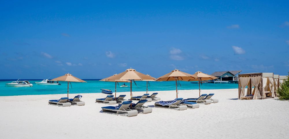 Sunlit beach with blue waters, beige umbrellas, and lounge chairs. Boats float on the horizon, and cabanas line the right side, evoking relaxation.
