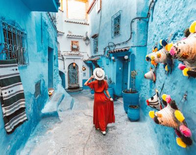 A woman in a red dress and hat walks down a narrow, vibrant blue alley in Chefchaouen, Morocco. Colorful hats adorn the walls, evoking a sense of wanderlust.