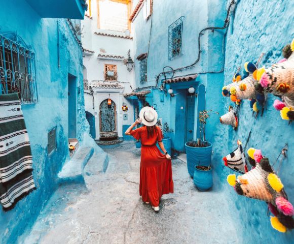 A woman in a red dress and hat walks down a narrow, vibrant blue alley in Chefchaouen, Morocco. Colorful hats adorn the walls, evoking a sense of wanderlust.