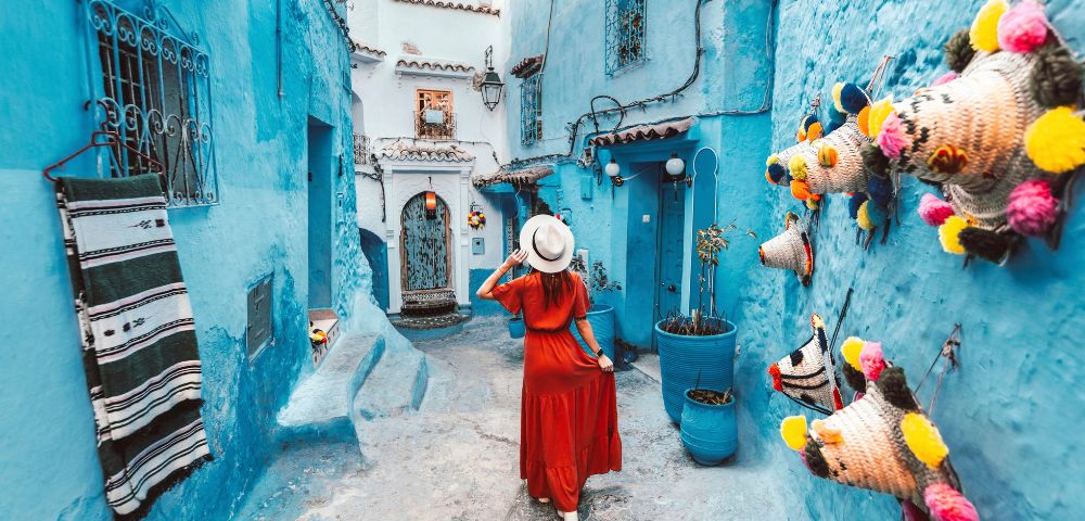 A woman in a red dress and hat walks down a narrow, vibrant blue alley in Chefchaouen, Morocco. Colorful hats adorn the walls, evoking a sense of wanderlust.