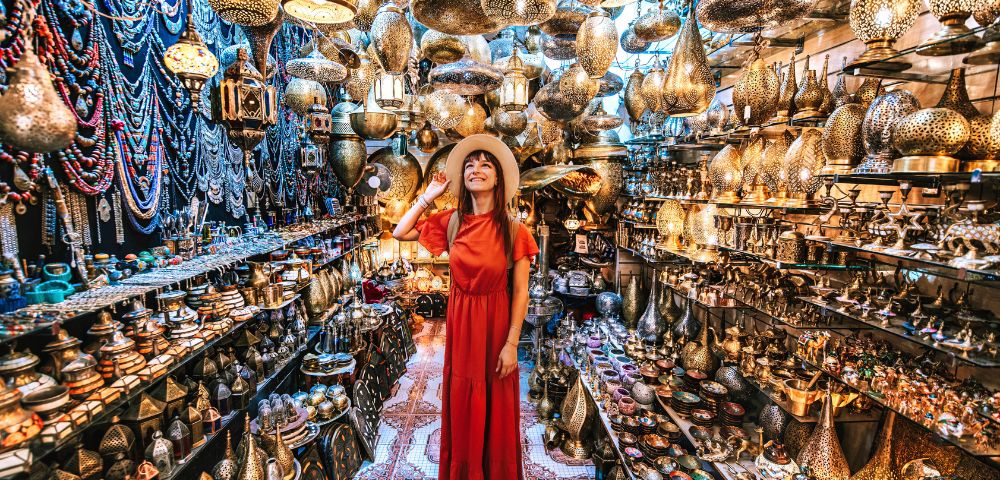 A woman in a red dress and hat stands smiling in a vibrant market stall filled with ornate hanging lanterns and pottery, exuding warmth and wonder.