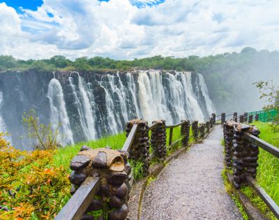 Pathway leading to a majestic waterfall with lush greenery on the sides, under a cloudy sky. The scene is vibrant and awe-inspiring.